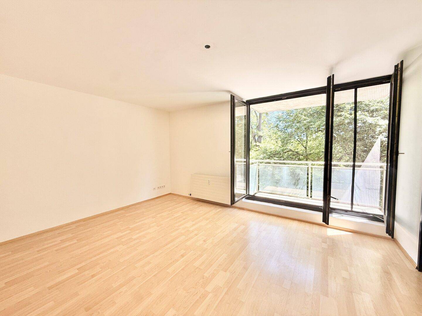 Helles, leeres Wohnzimmer mit Parkettboden und großen Fenstern zum Balkon.