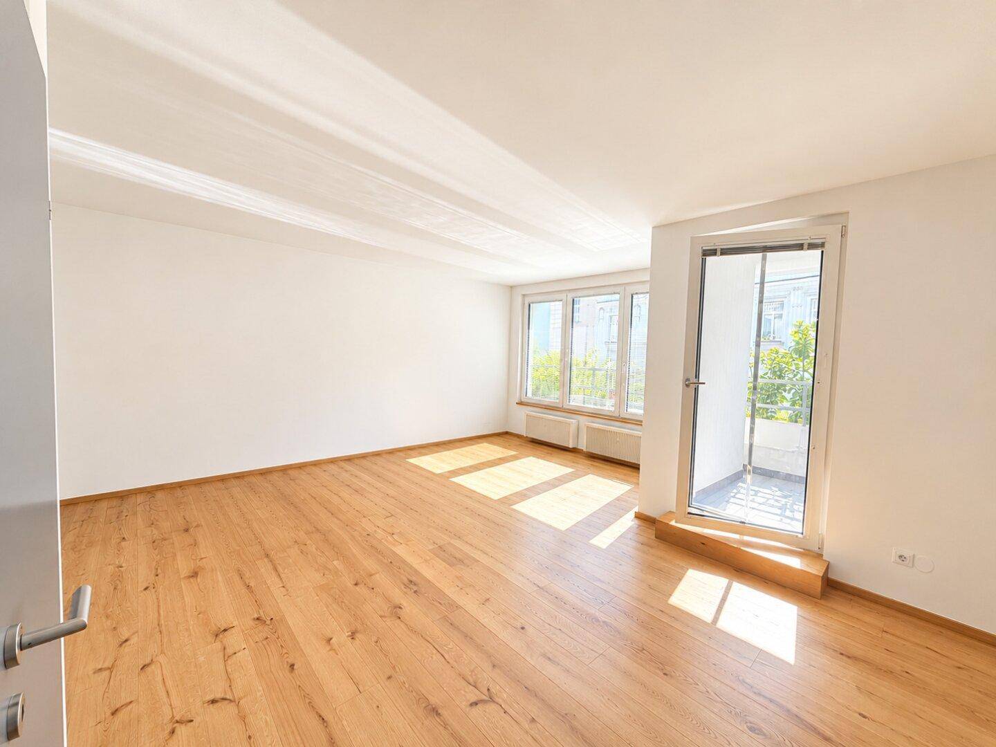 Helles Wohnzimmer mit Holzboden und großen Fenstern, die viel Tageslicht hereinlassen.