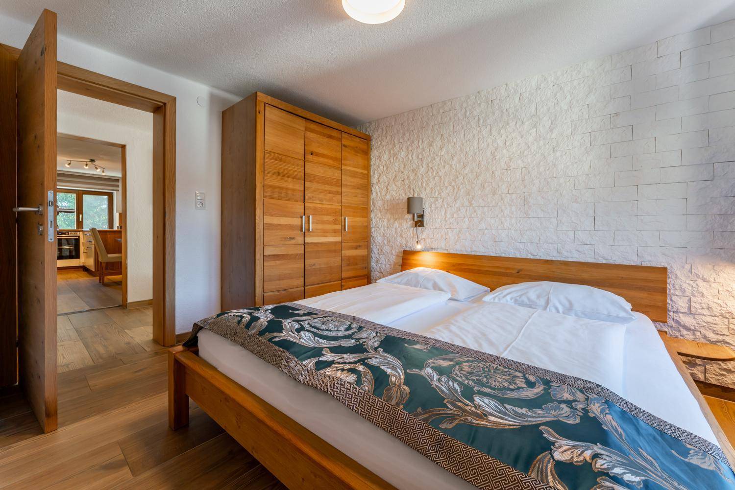Helles Schlafzimmer mit Doppelbett, Holzschrank und strukturierter Wand, verbunden mit dem Wohnbereich.