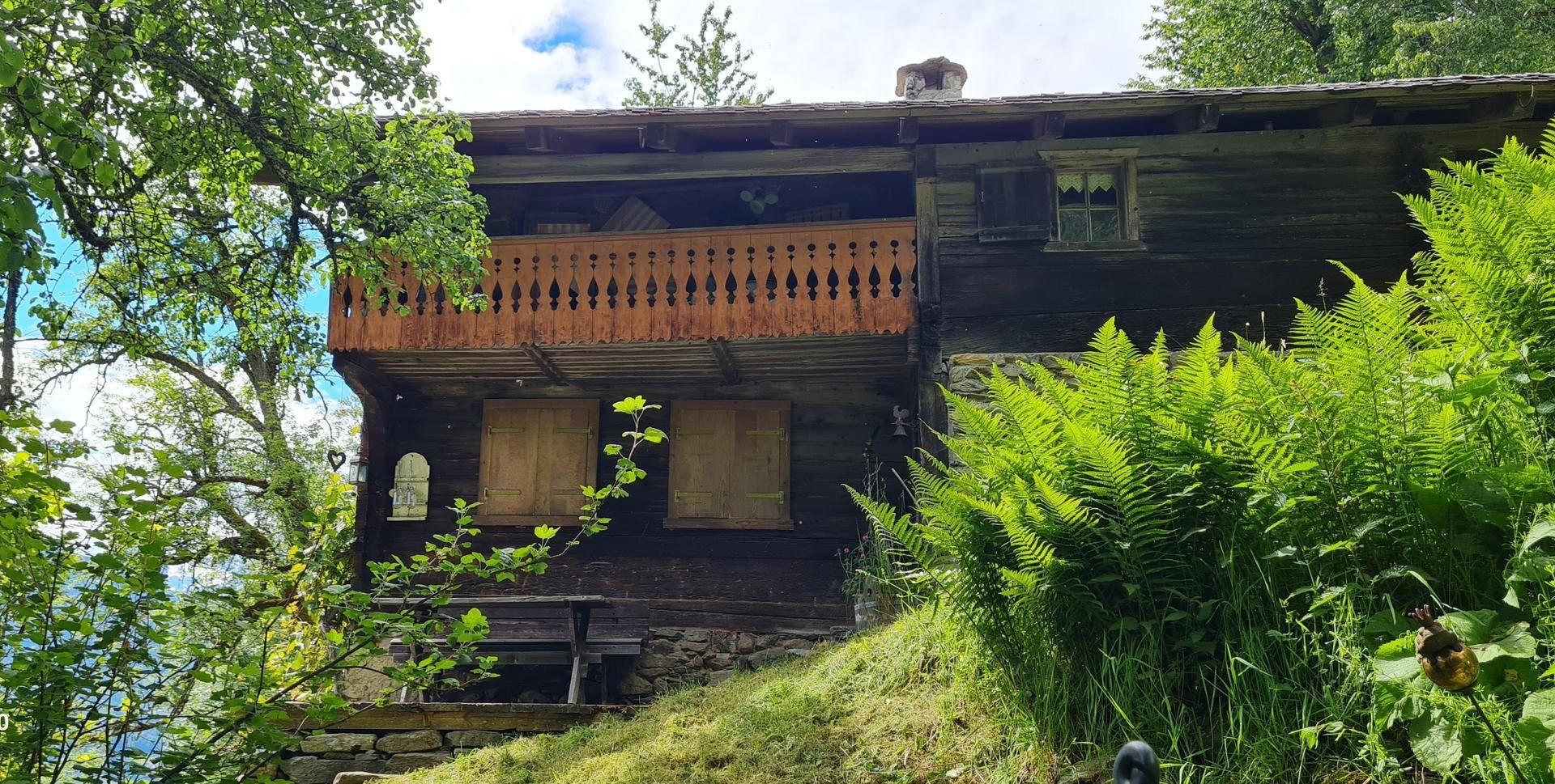 Traditionelles Holzhaus mit geschnitztem Holzbalkon und üppigem Grün im Vordergrund.
