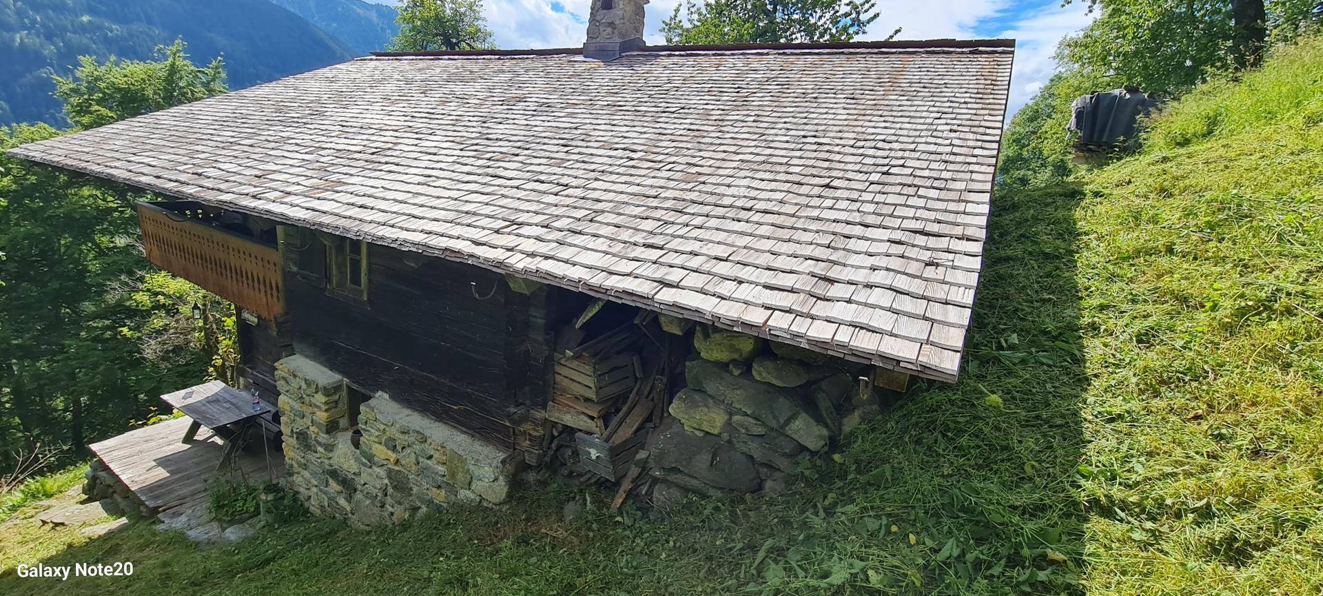 Seitenansicht eines traditionellen Holzhauses mit Schindeldach und Steinfundament in grüner Umgebung.