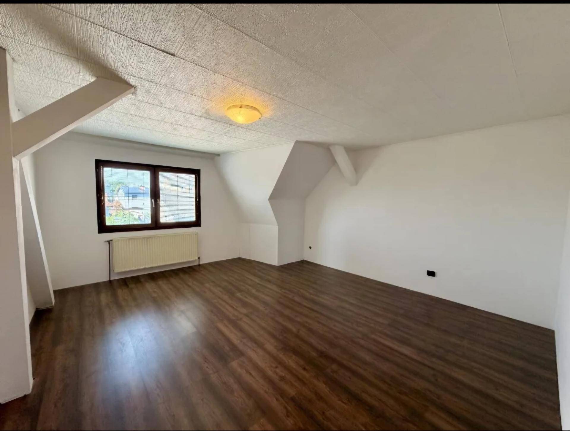 Geräumiges Dachzimmer mit Laminatboden, weiß gestrichenen Wänden und Heizkörper unter dem Fenster.