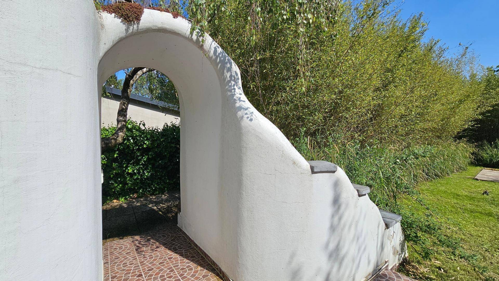 Weißer Torbogen in einem Garten mit gepflastertem Bereich und üppigem Grün im Hintergrund.