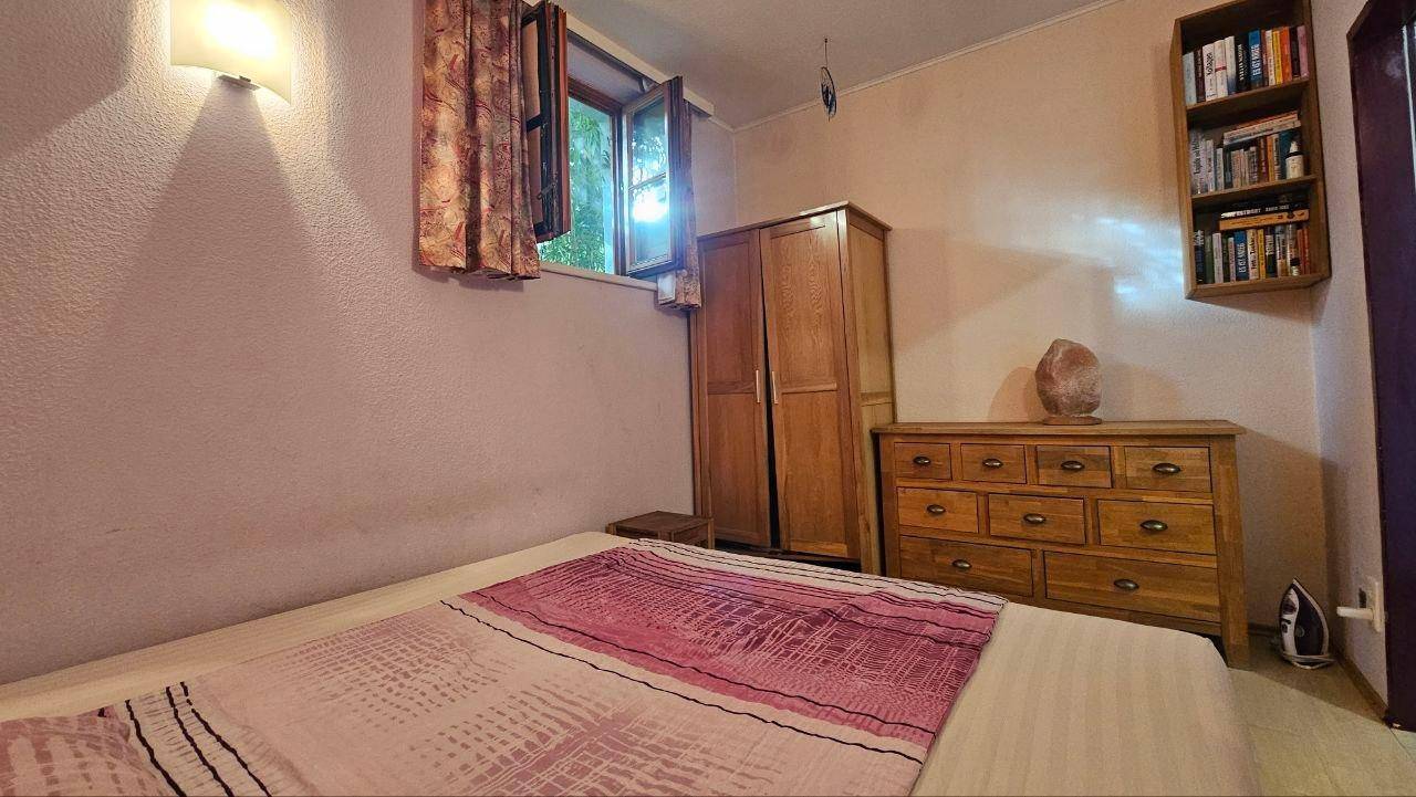 Schlafzimmer mit Holzkleiderschrank, Kommode und einem geöffneten Fenster, das Licht hereinlässt.