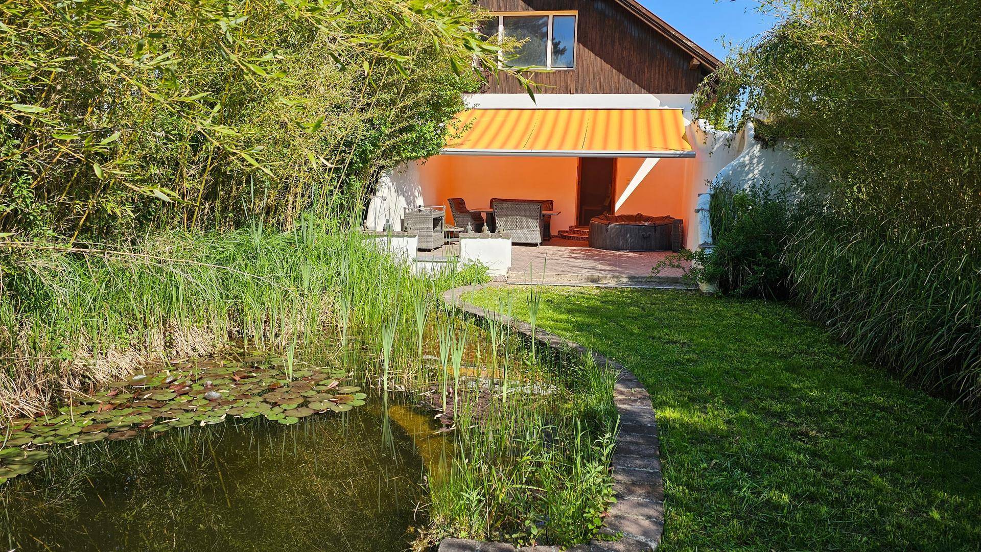 Idyllischer Garten mit Teich, üppiger Vegetation und einer Terrasse mit Whirlpool und Gartenmöbeln.