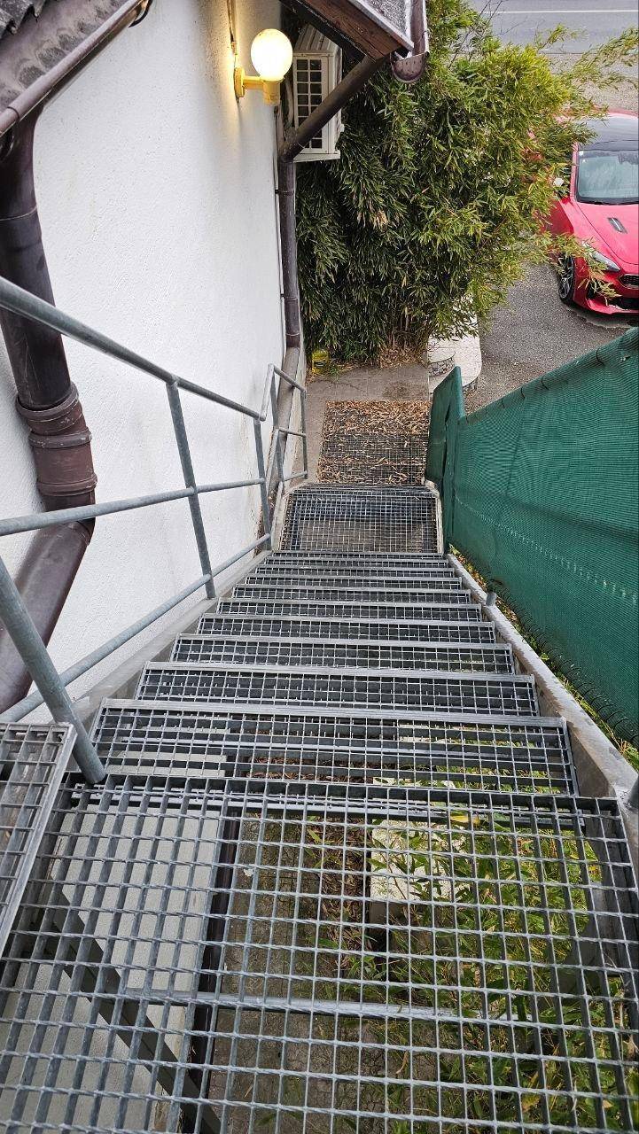 Metalltreppe führt vom Gebäude nach unten, umgeben von Grün und einem roten Auto im Hintergrund.