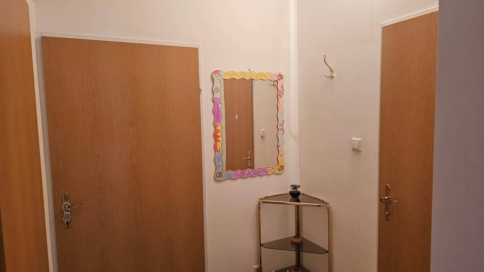 Kleiner Flur mit zwei Holztüren, einem bunten Spiegel und einer Eckablage, einfach eingerichtet.