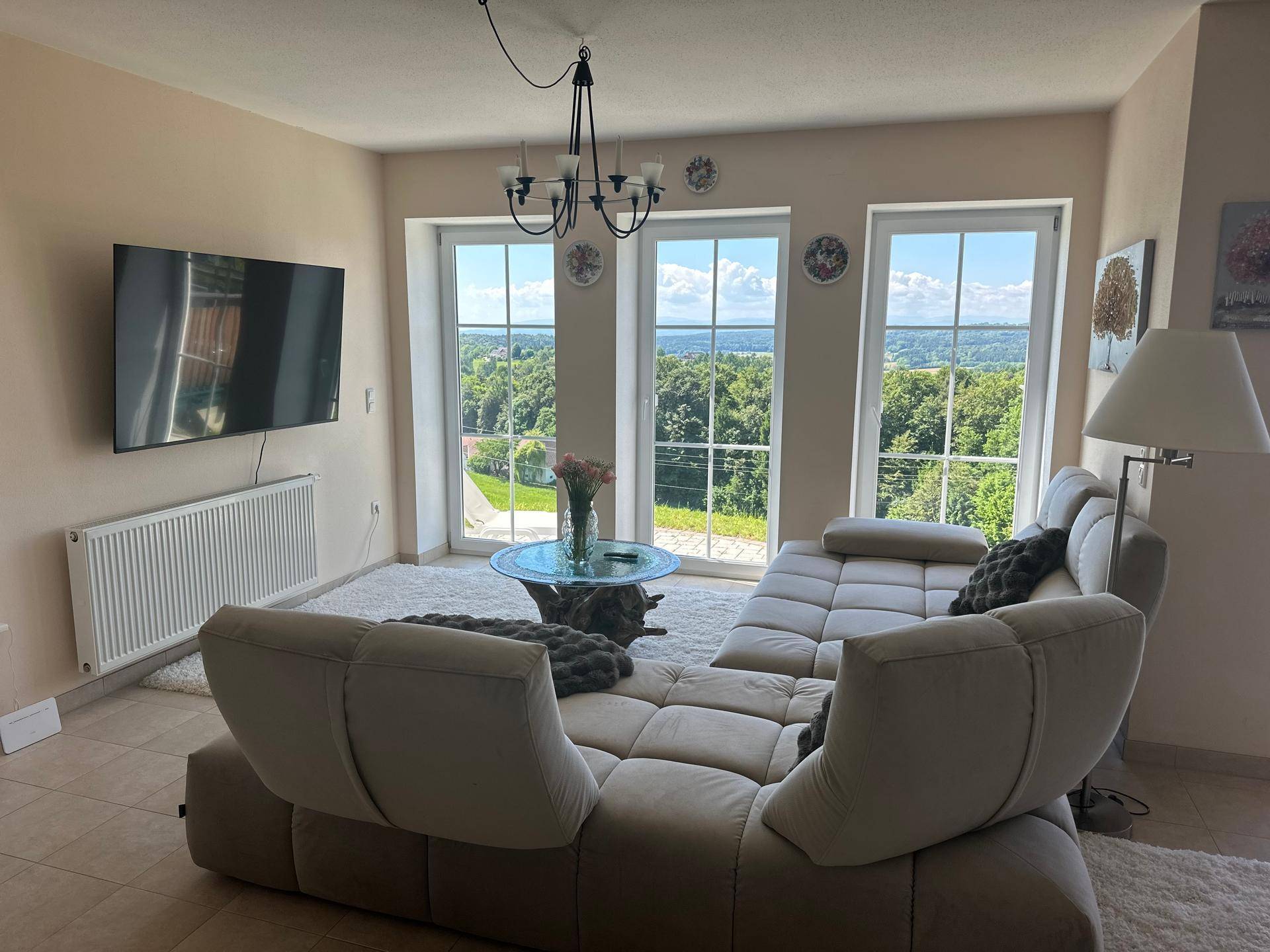 Helles Wohnzimmer mit beigem Sofa, Fliesenboden und weitem Blick ins Grüne.