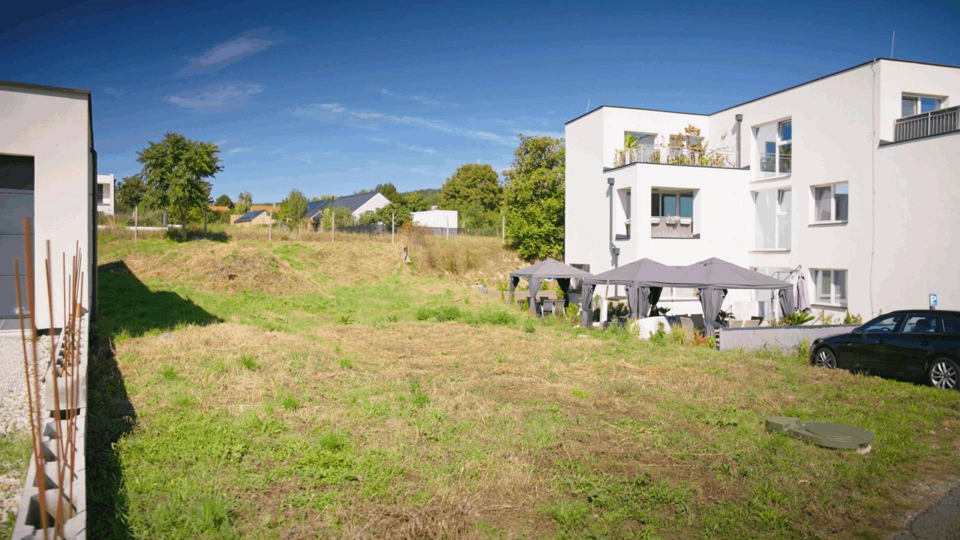Unbebautes Grundstück neben einem modernen Mehrfamilienhaus mit möblierten Outdoor-Sitzbereichen.