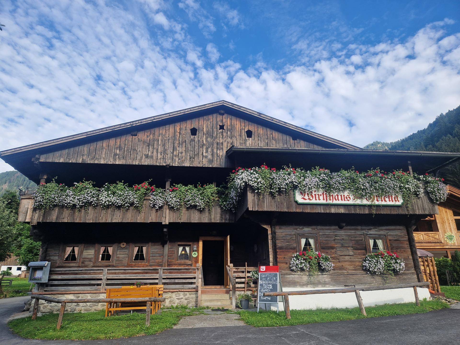 Traditionelles Holzgebäude eines Wirtshauses mit Blumenkästen und Bergblick unter blauem Himmel.