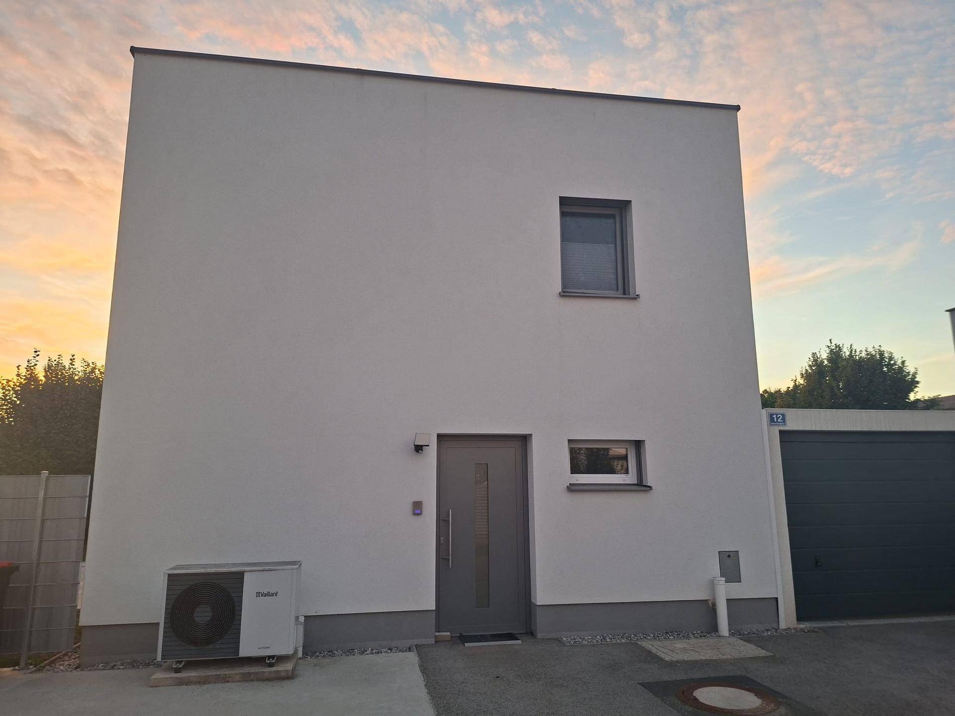 Vorderansicht des Hauses mit Eingangstür, Fenster und installierter Wärmepumpe.