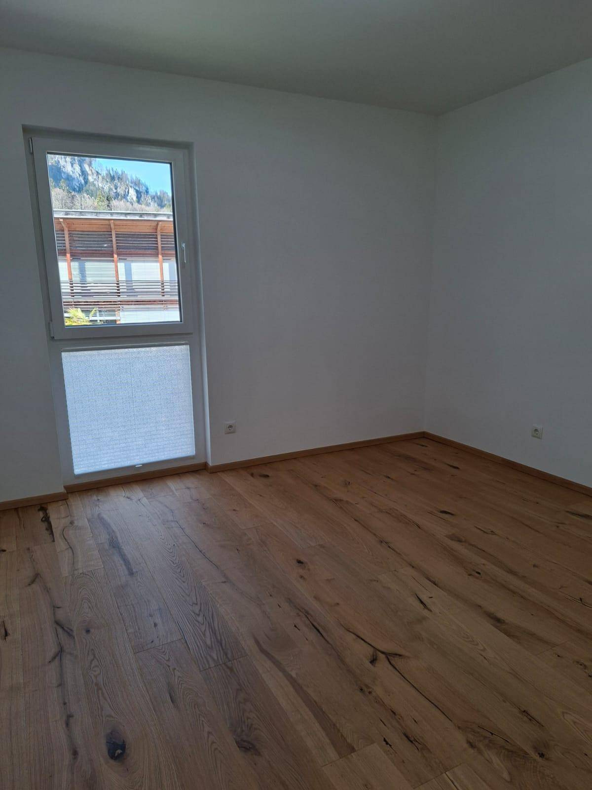 Leeres Zimmer mit hellem Parkettboden und Fenster mit Blick auf die Berge.