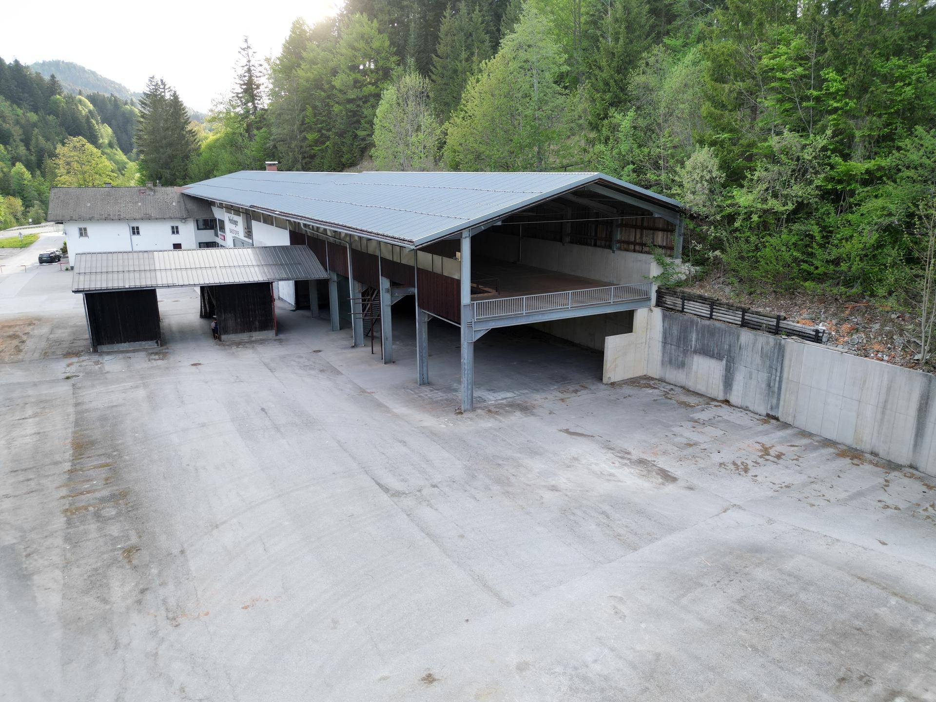 Rückansicht des Gewerbegebäudes mit großer Betonfläche, offener Halle und angrenzendem Waldgebiet.
