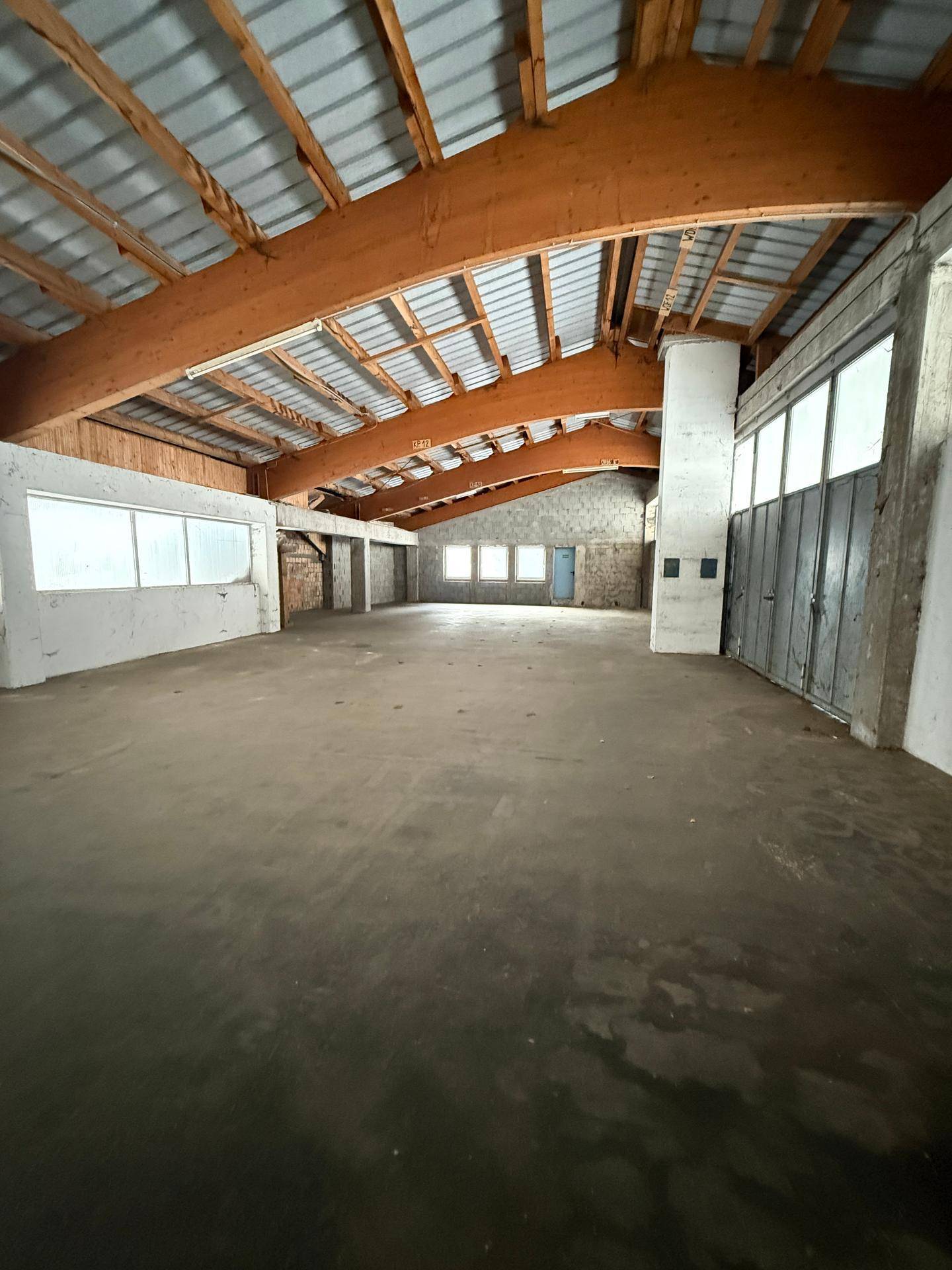 Geräumige Lagerhalle mit hoher Holzkonstruktion, Betonboden und großen Fenstern, die viel Tageslicht hereinlassen.