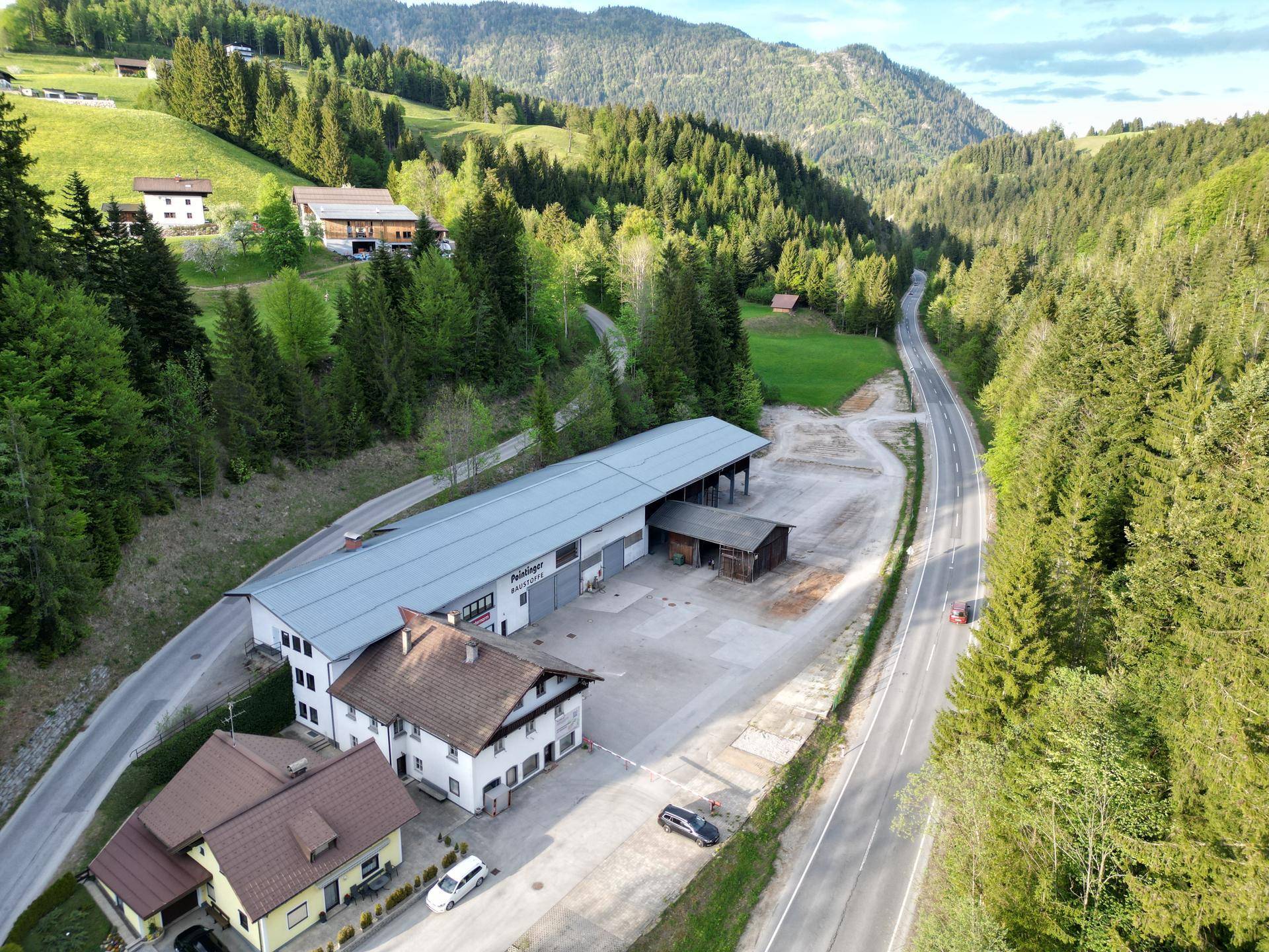 Weitläufige Luftaufnahme des Gewerbeobjekts an einer Straße, eingebettet in eine grüne Hügellandschaft.