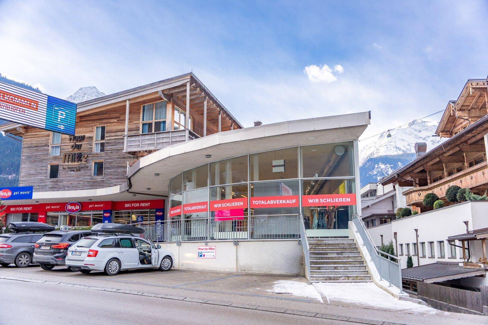 Geschäftshaus mit moderner Architektur, großen Glasflächen und Parkplätzen, umgeben von alpiner Landschaft.