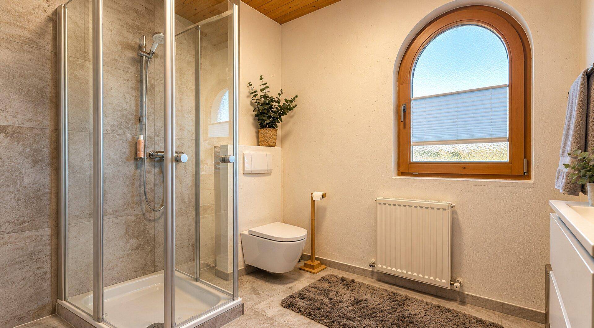Badezimmer mit Duschkabine, wandhängendem WC, Heizkörper und Rundbogenfenster.
