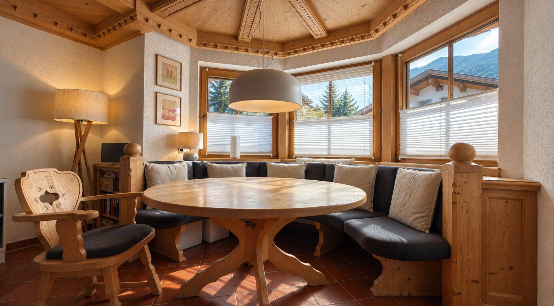 Heller Essbereich mit runder Holztafel und Eckbank, umgeben von Fenstern mit Bergblick.