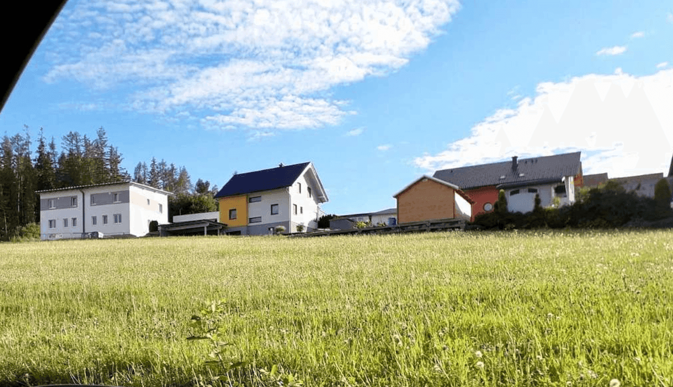 Panoramablick über eine grüne Wiese mit modernen Wohnhäusern und Wald am Horizont.