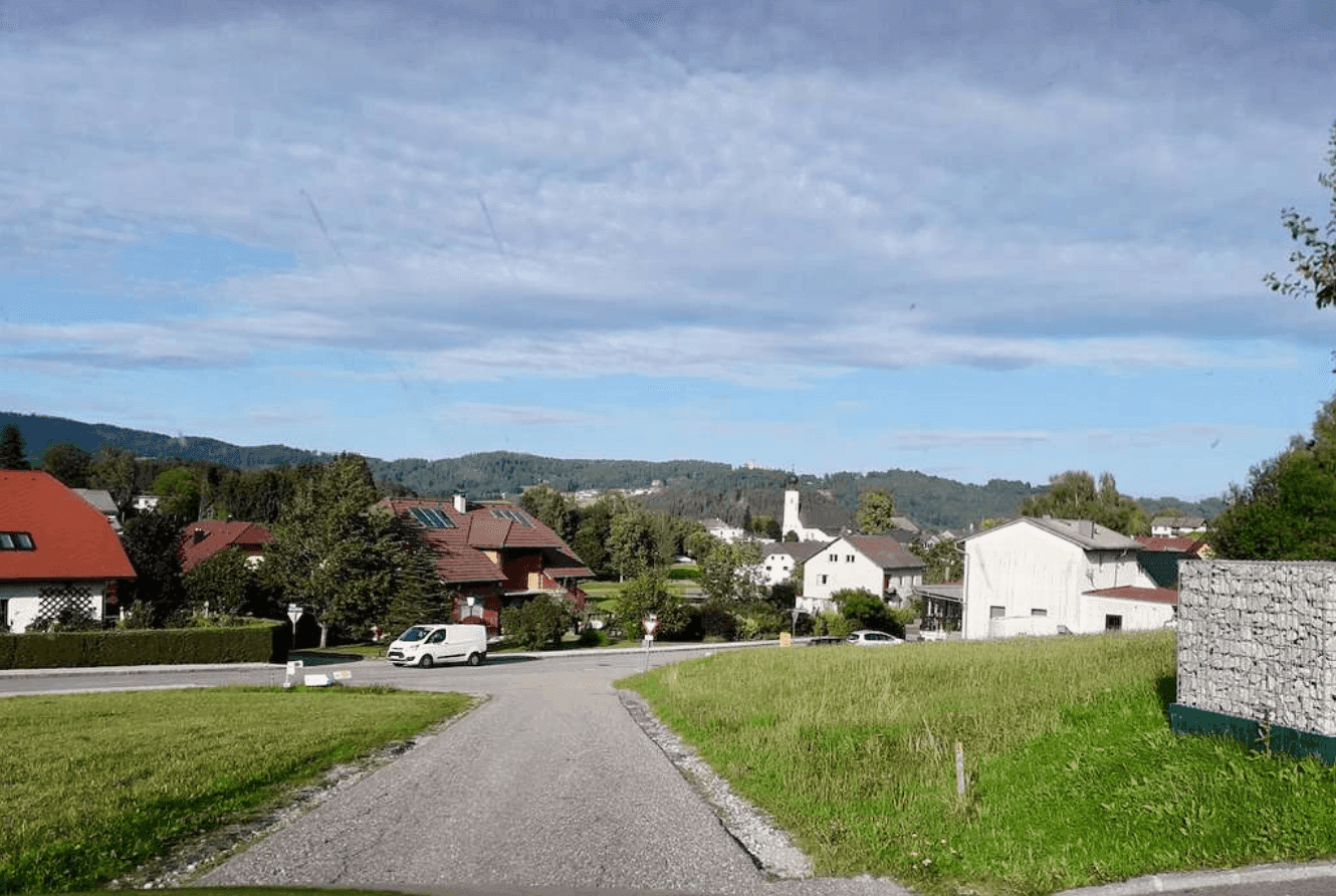 Blick auf eine ländliche Siedlung mit Häusern, Bäumen und einem Kirchturm in der Ferne.