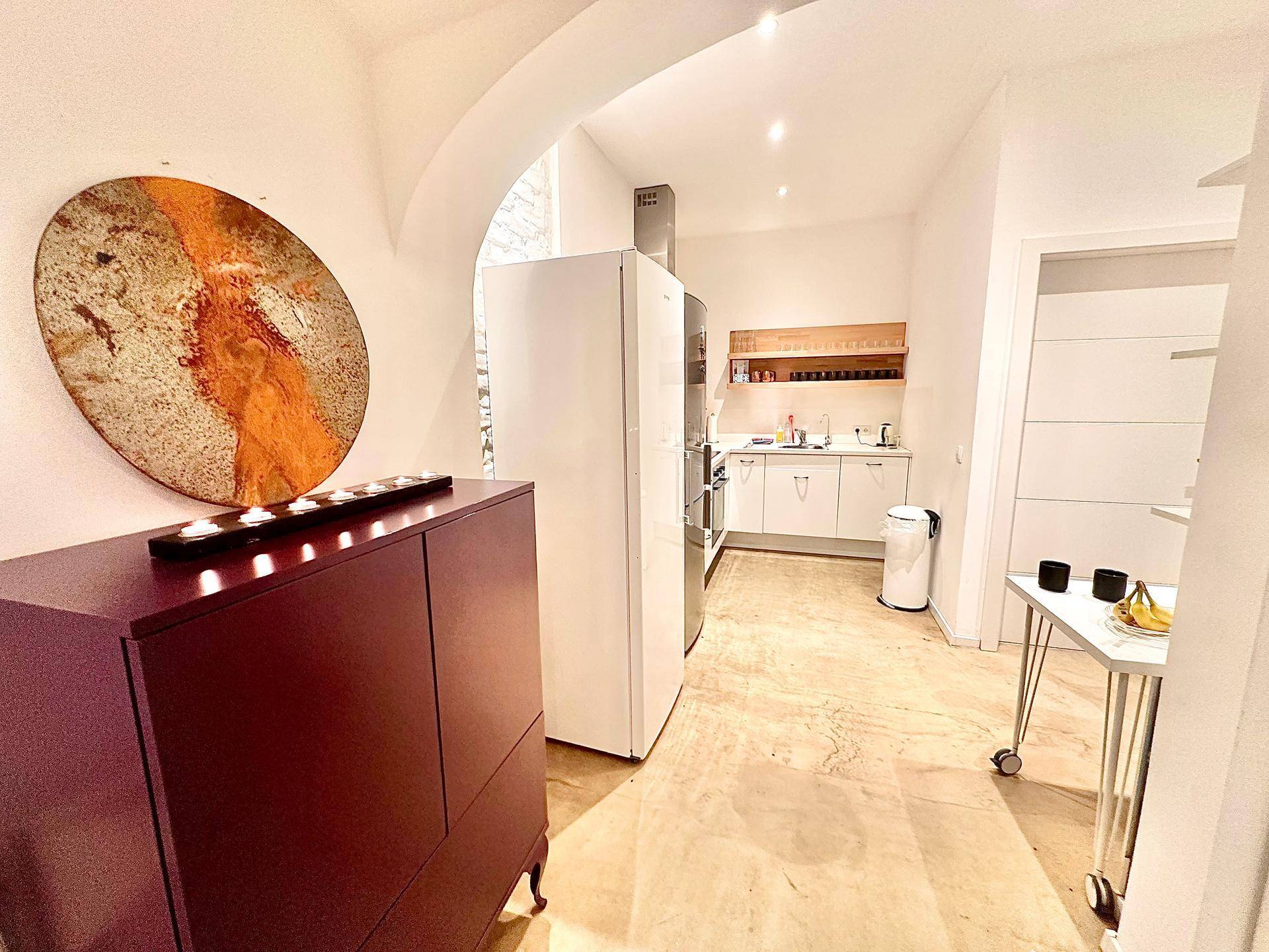 Funktionale Küche mit weißen Fronten, offenem Regal und großem Kühlschrank, verbunden mit einem Flurbereich.