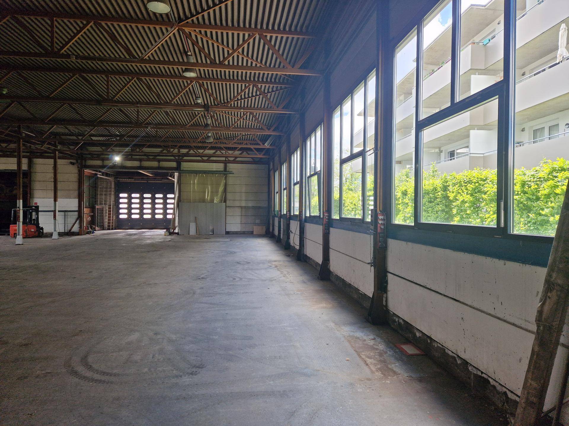 Lange Lagerhalle mit hohen Decken, großen Fenstern entlang einer Wand und Blick auf ein angrenzendes Gebäude.