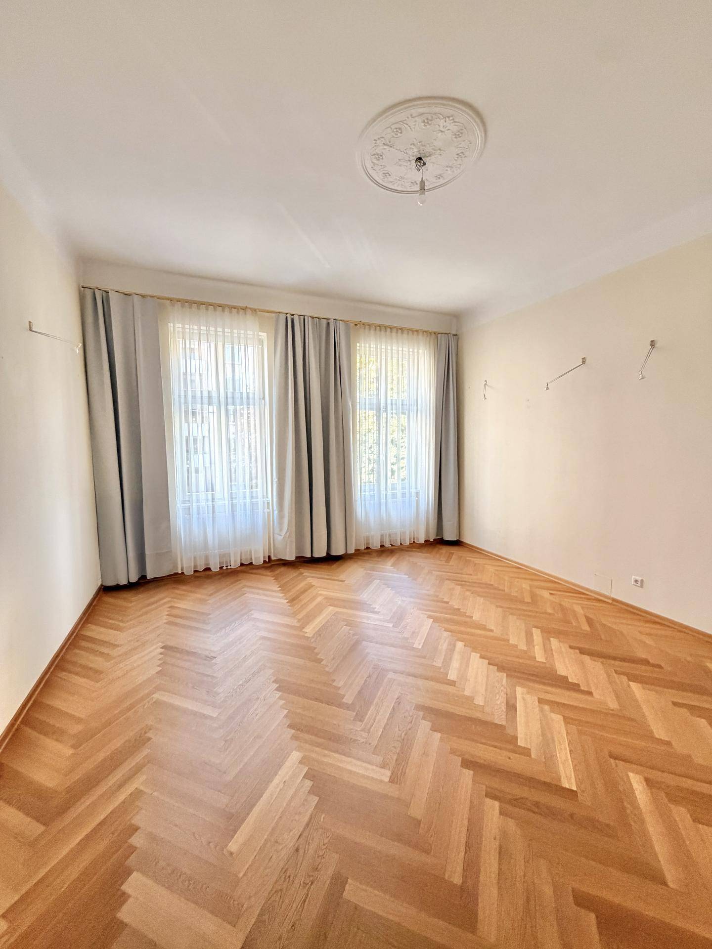 Geräumiges Zimmer mit klassischem Fischgrätparkett, hohen Decken und dekorativer Stuckrosette.