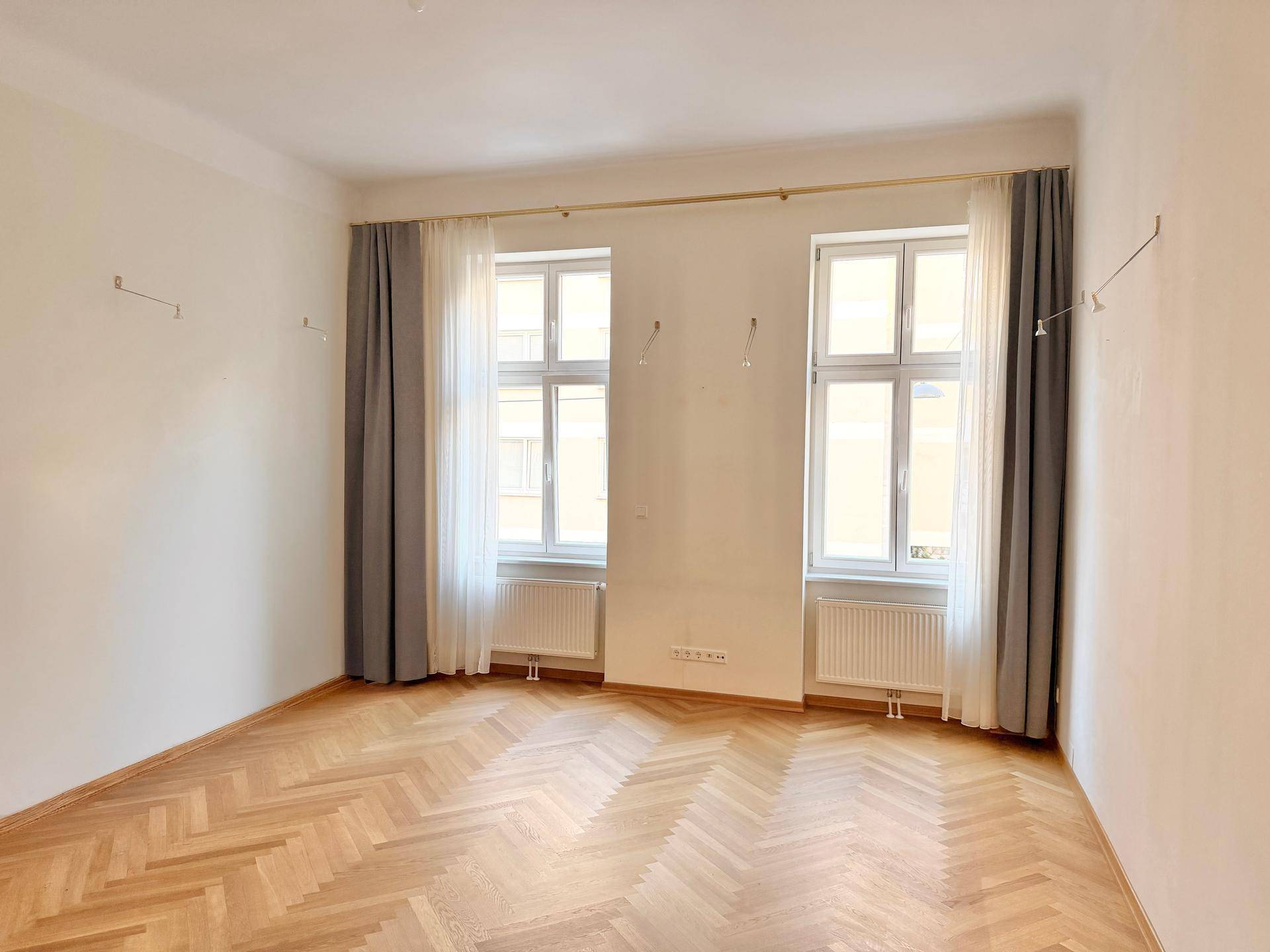 Geräumiges Zimmer mit elegantem Fischgrätparkett, zwei großen Fenstern und Heizkörpern darunter.