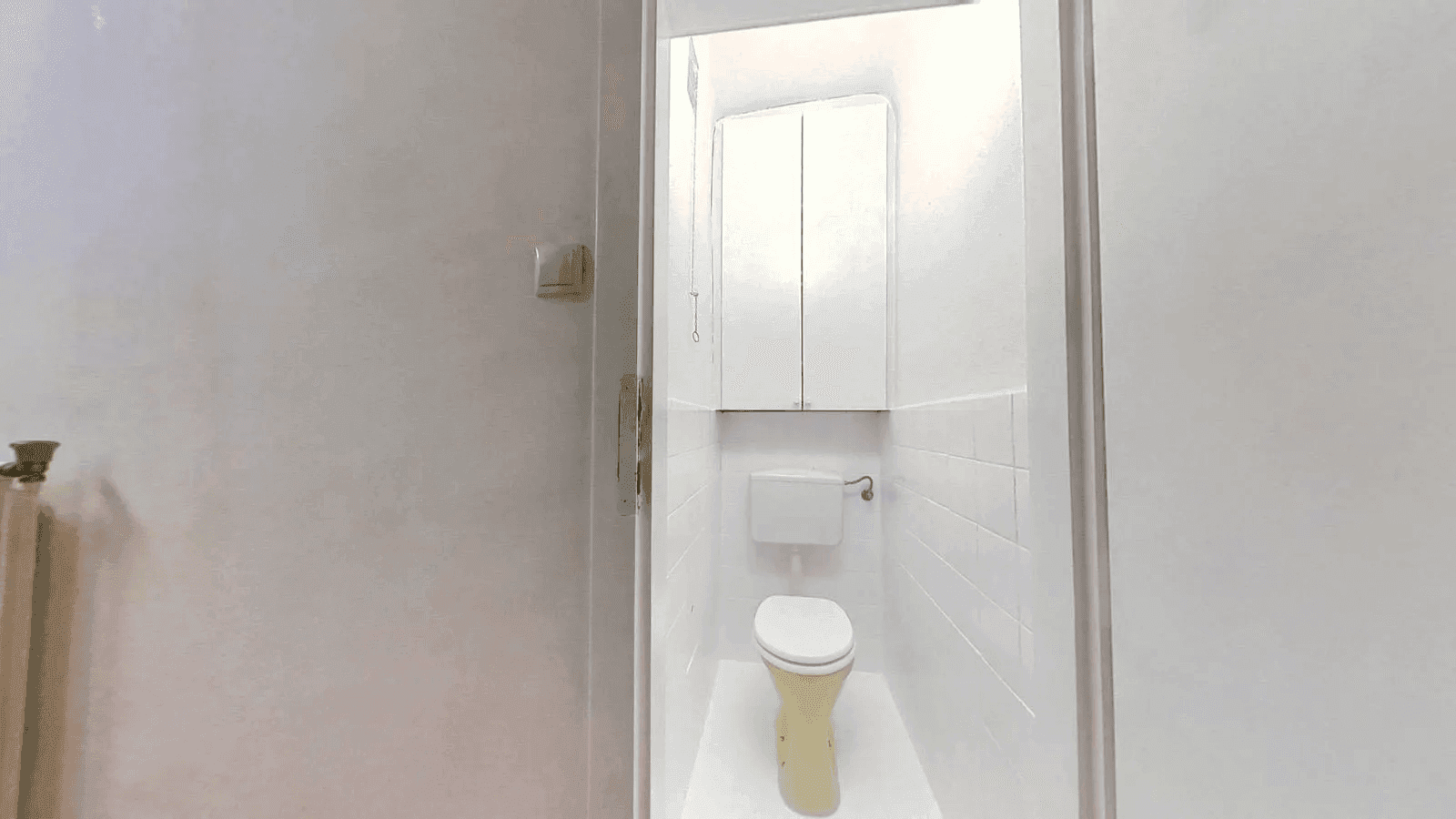 Kleines separates WC mit weiß gefliesten Wänden und einem Spülkasten über der Toilette.