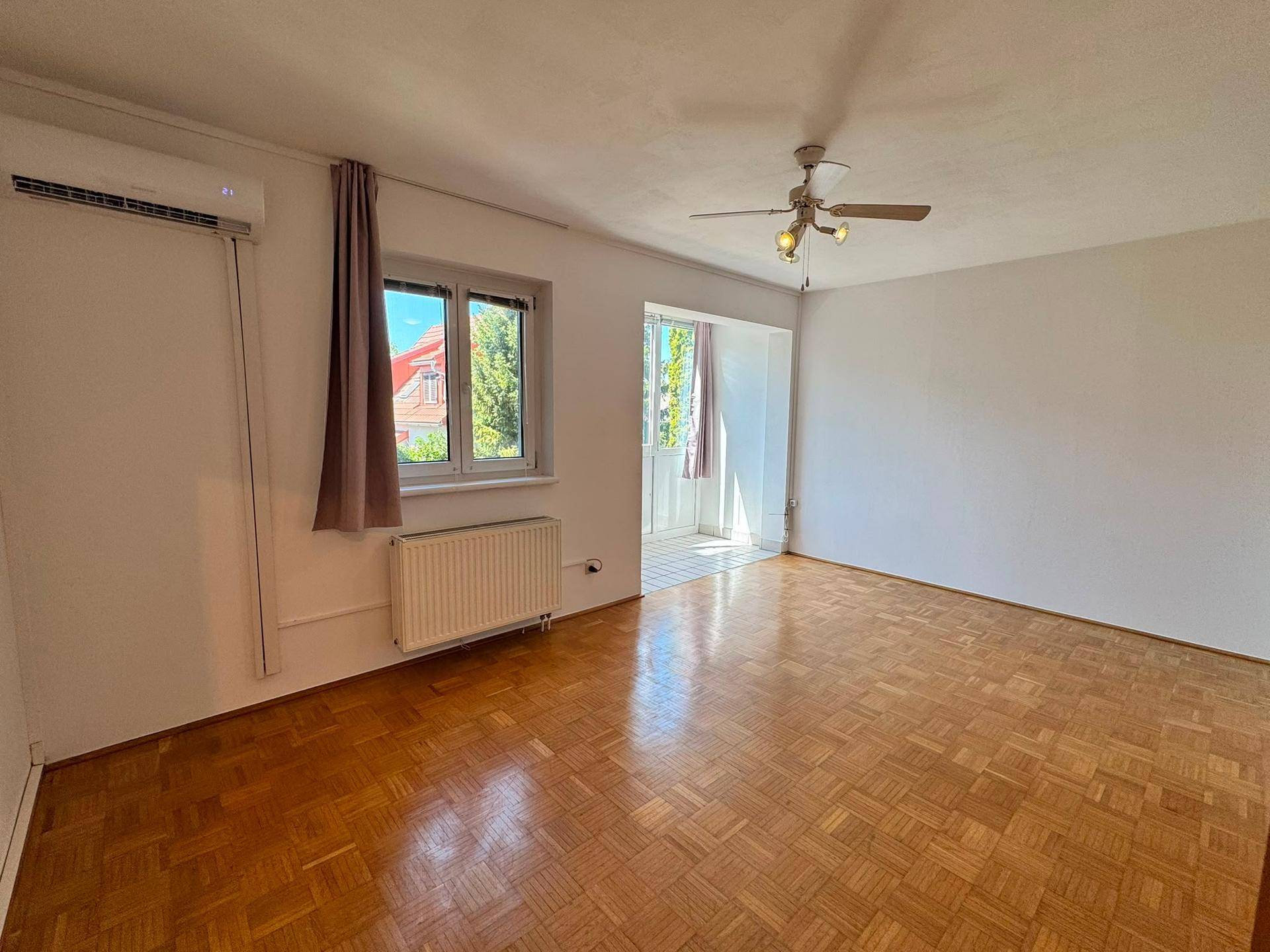 Geräumiges Wohnzimmer mit Parkettboden, Fenster und Zugang zum Balkon.