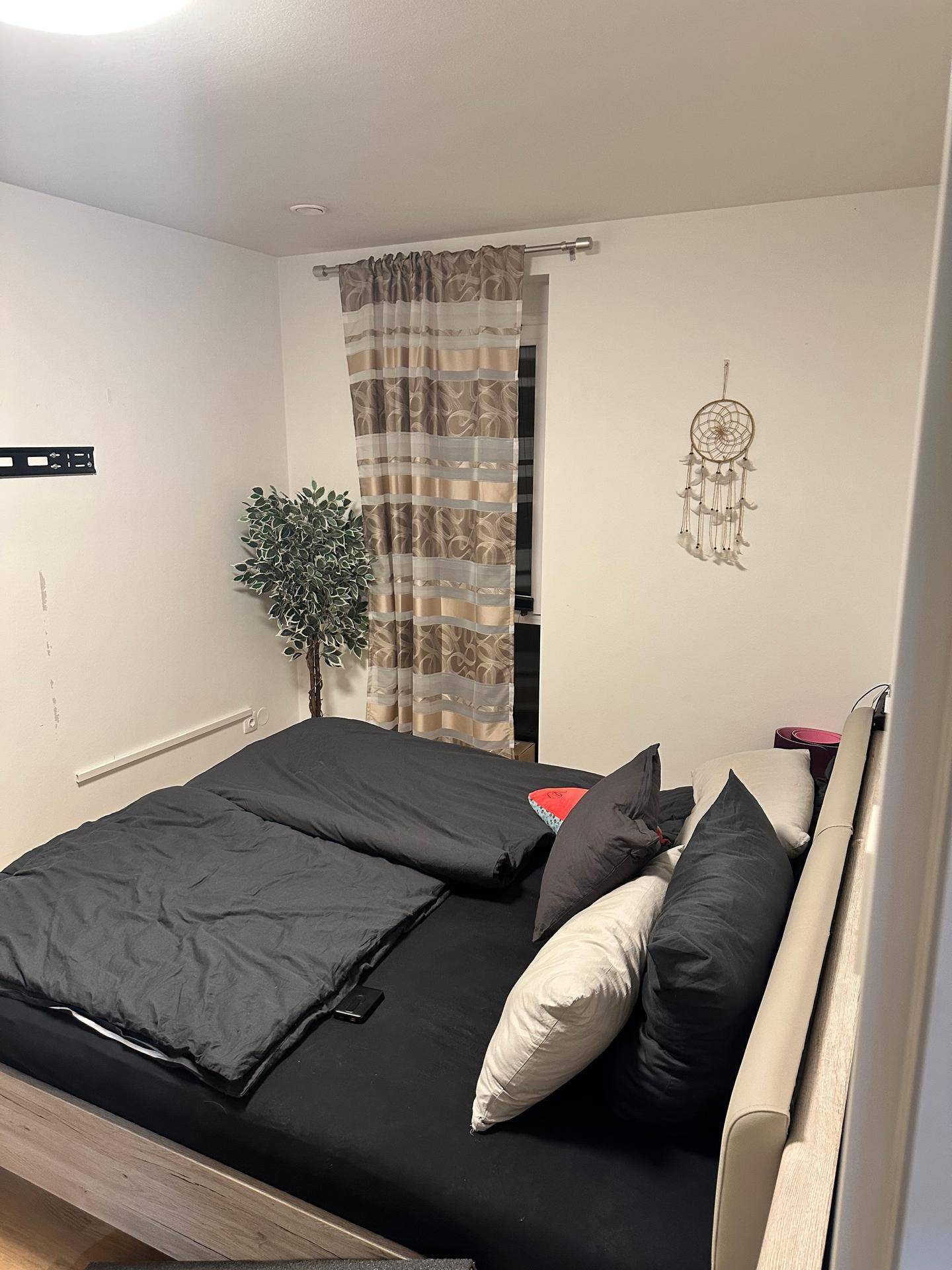 Schlafzimmer mit Doppelbett, schlichten Vorhängen und einem dekorativen Traumfänger an der Wand.
