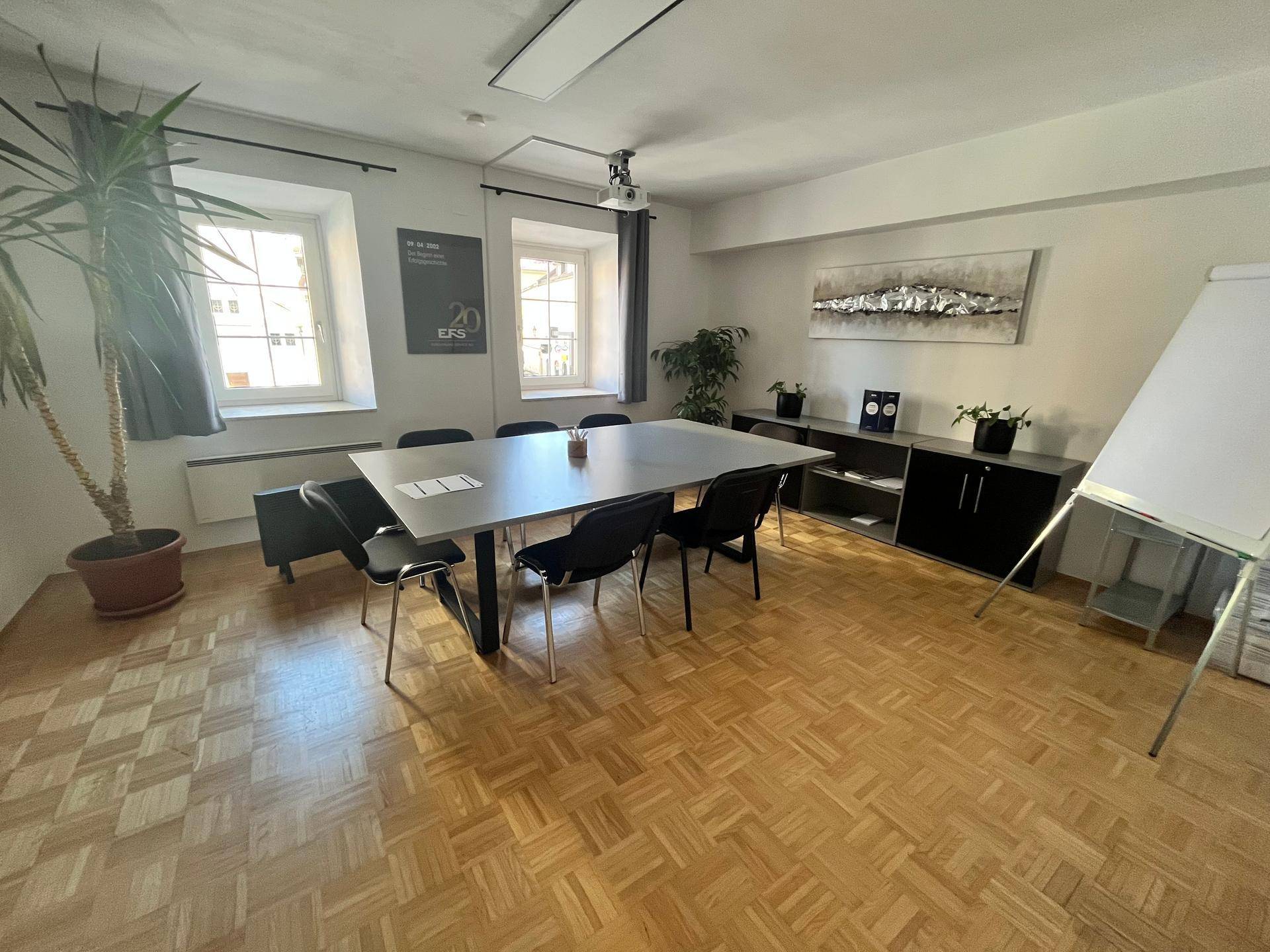 Großer Konferenzraum mit Holzboden, langem Tisch, Stühlen und Fenstern mit Tageslicht, ideal für Besprechungen.