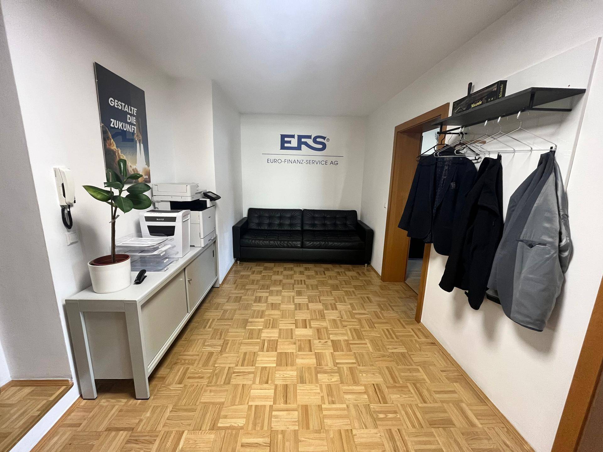Empfangsbereich mit Parkettboden, schwarzem Sofa und einem Schrank mit Drucker, hell beleuchtet.