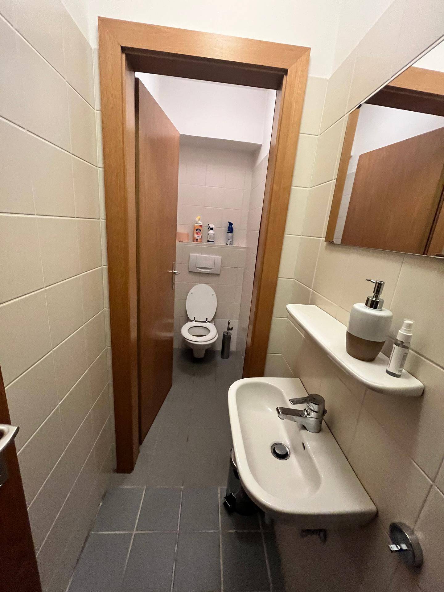 Kleine Gästetoilette mit Waschbecken, Spiegel und weißen Fliesen an Wänden und Boden, sauber und funktional.