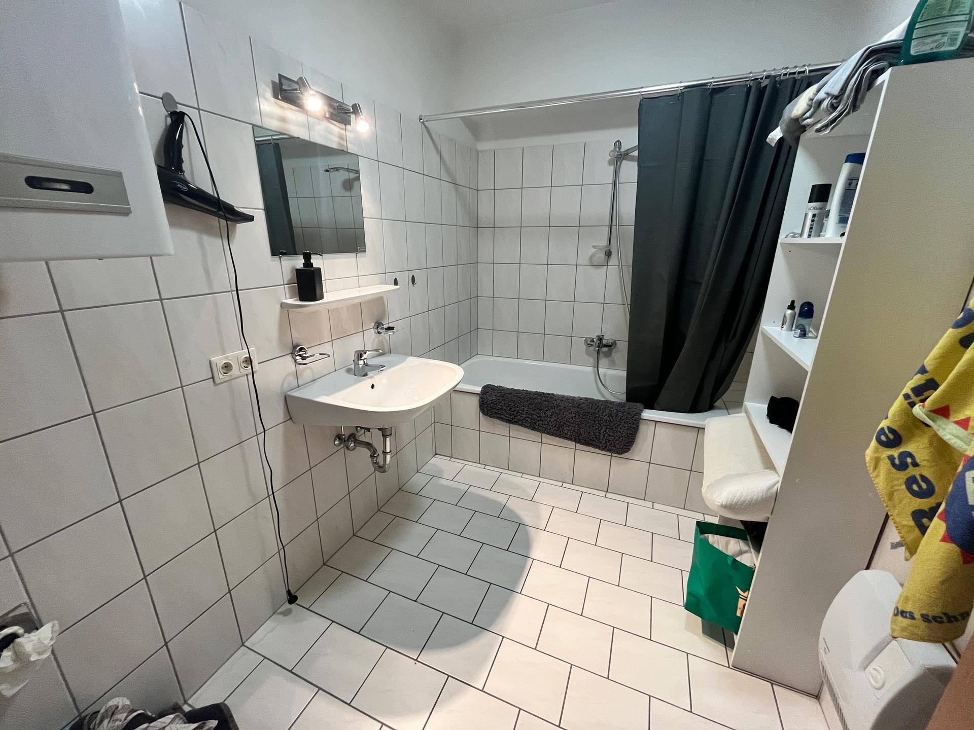 Badezimmer mit Badewanne, Duschvorhang, Waschbecken und weißen Wand- und Bodenfliesen, funktional eingerichtet.