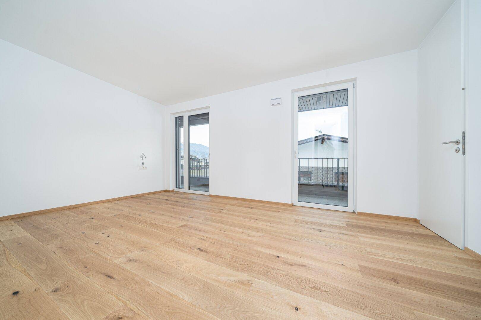 Leeres, helles Zimmer mit Holzboden und zwei Fenstertüren, die zu einem Balkon führen.