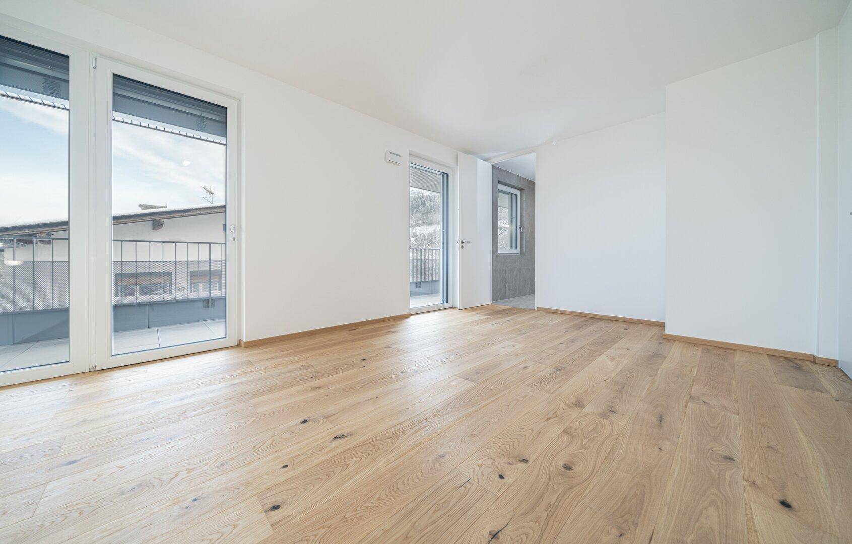 Helles, leeres Zimmer mit Holzboden und zwei Türen, die zu einem Balkon führen.