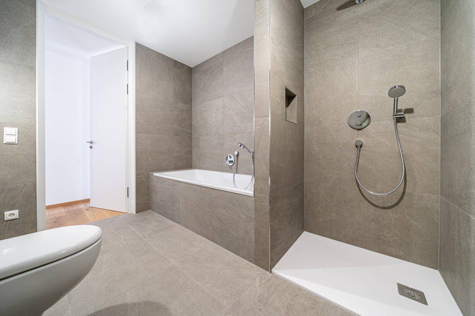 Modernes Badezimmer mit grauen Fliesen, Badewanne, ebenerdiger Dusche und Toilette.