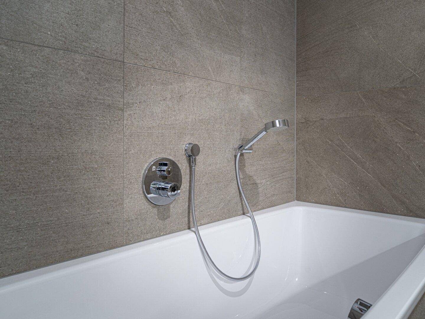 Nahaufnahme der Badewannenarmatur mit Handbrause in einem modern gefliesten Badezimmer.
