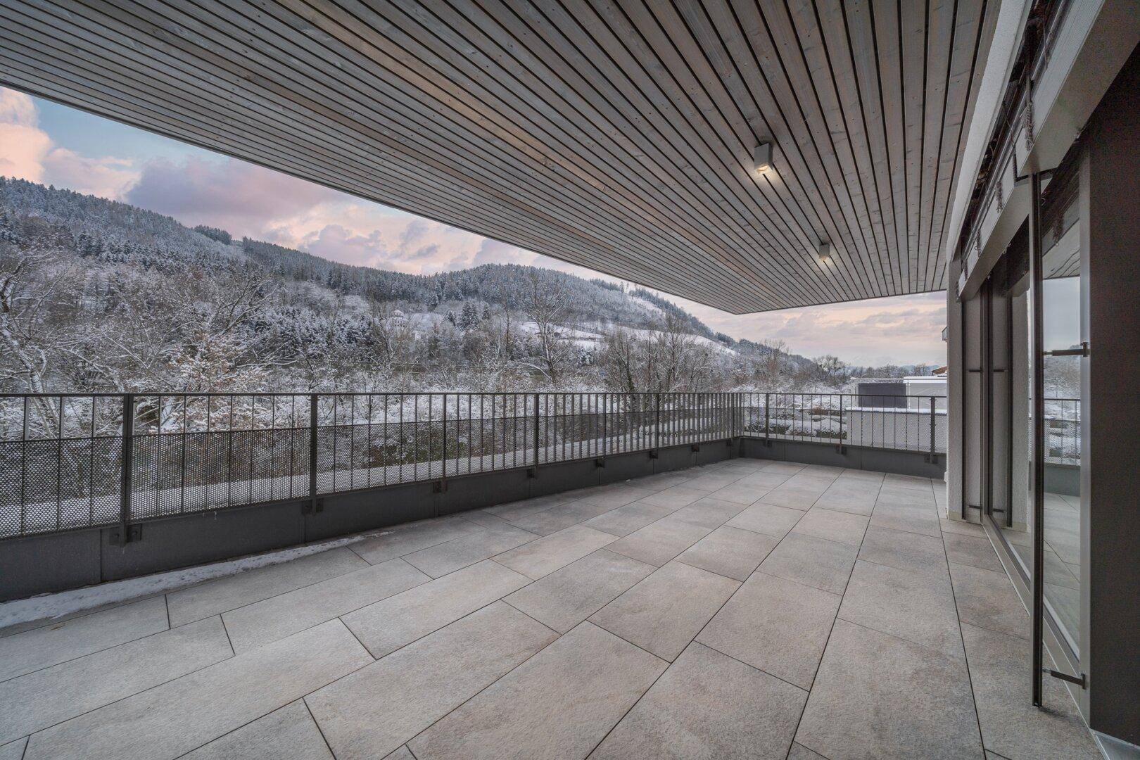 Große, geflieste Terrasse mit Holzdecke und weitem Blick auf die verschneite Berglandschaft im Winter.
