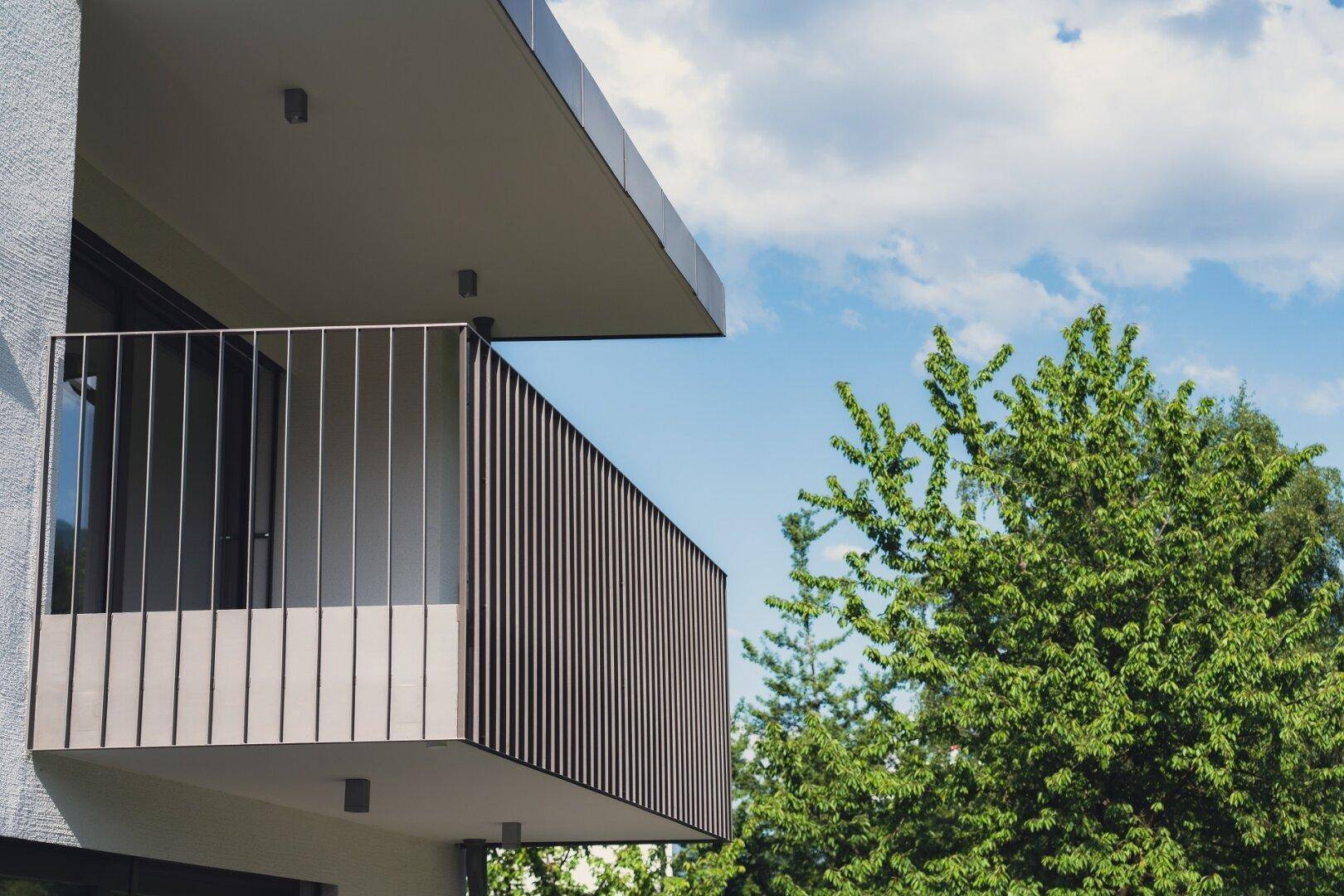 Moderner Balkon mit vertikalen Geländerstäben und Blick ins Grüne, unter blauem Himmel.