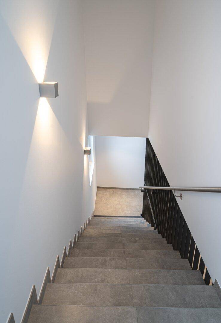 Modernes Treppenhaus mit grauen Fliesenstufen, weißen Wänden und stilvoller Wandbeleuchtung.
