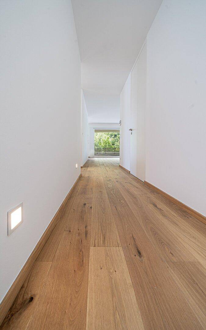 Langer, heller Flur mit Holzboden und integrierten Wandleuchten, der zu einem Fenster führt.