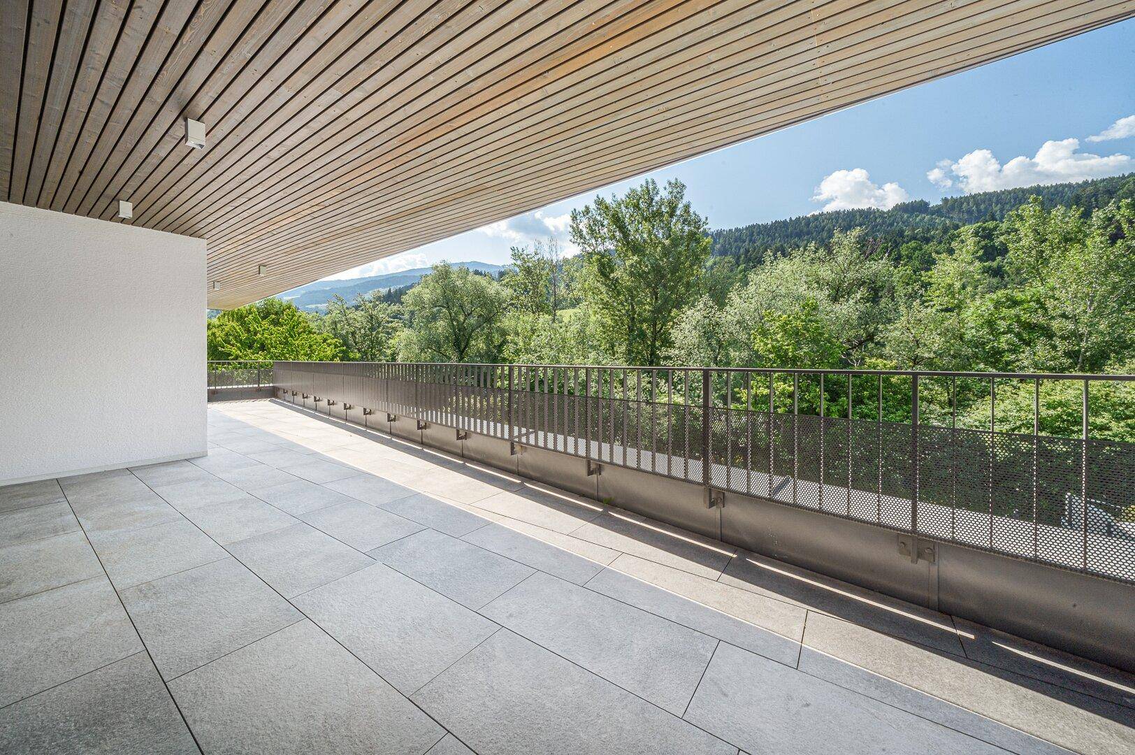Sonniger Terrassenbereich mit Fliesenboden, Metallgeländer und weitem Blick auf bewaldete Hügel.