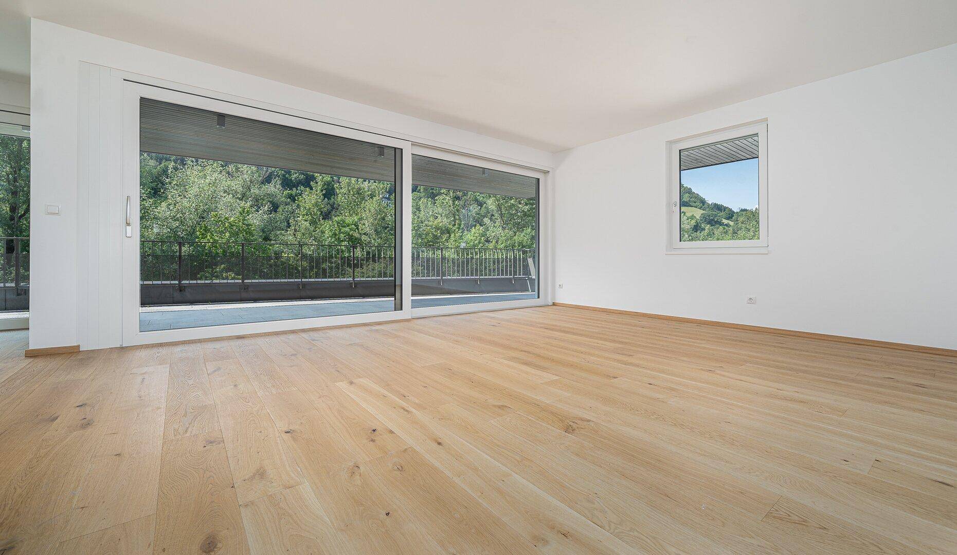 Leeres, helles Wohnzimmer mit Holzboden, großen Fenstern und Blick auf die grüne Landschaft.