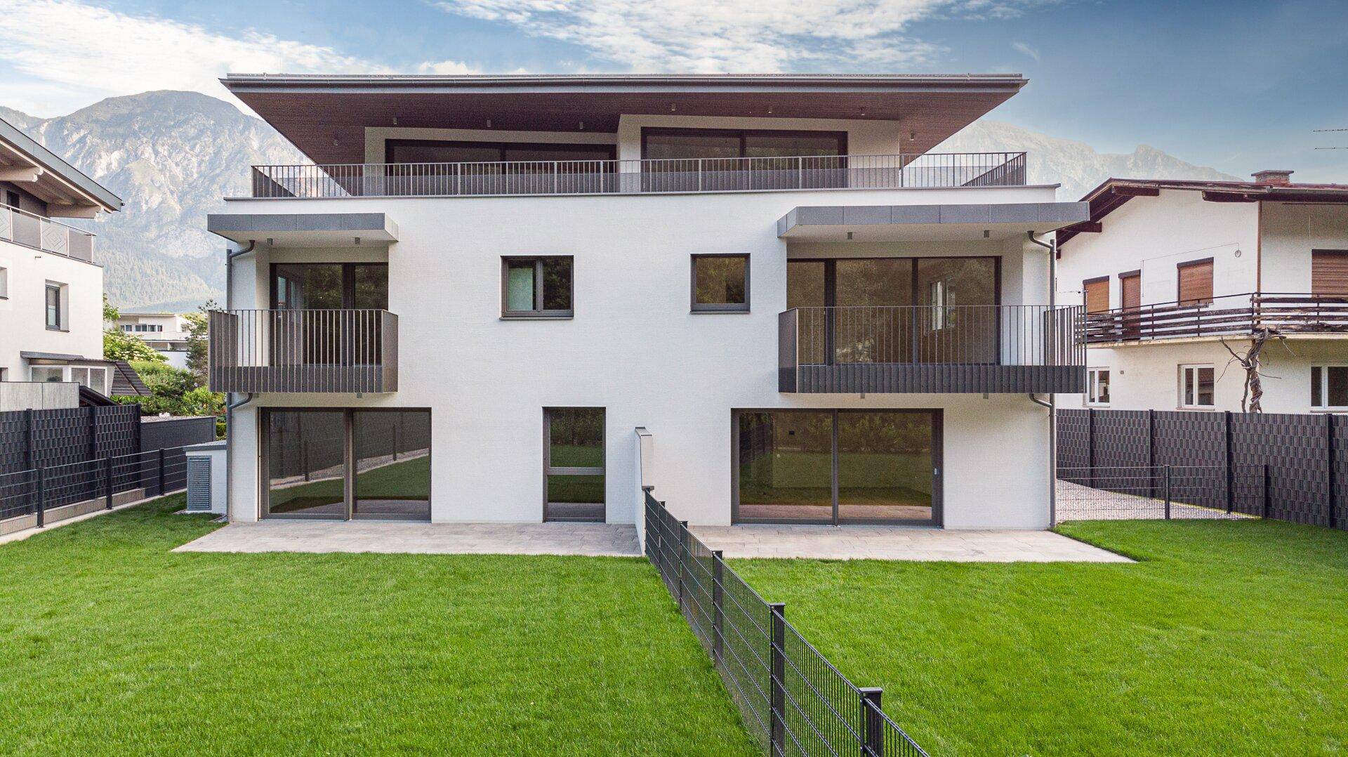 Moderne Wohnanlage mit weißer Fassade, Balkonen und gepflegtem Rasen vor alpiner Bergkulisse.