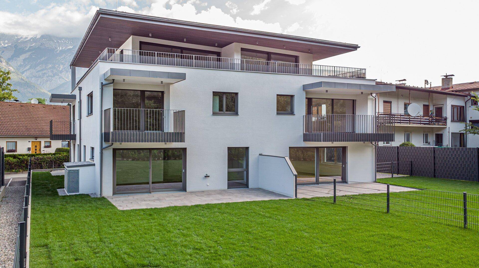Neubau-Wohngebäude mit weißer Putzfassade, großen Fenstern und Zugang zu einer Rasenfläche.