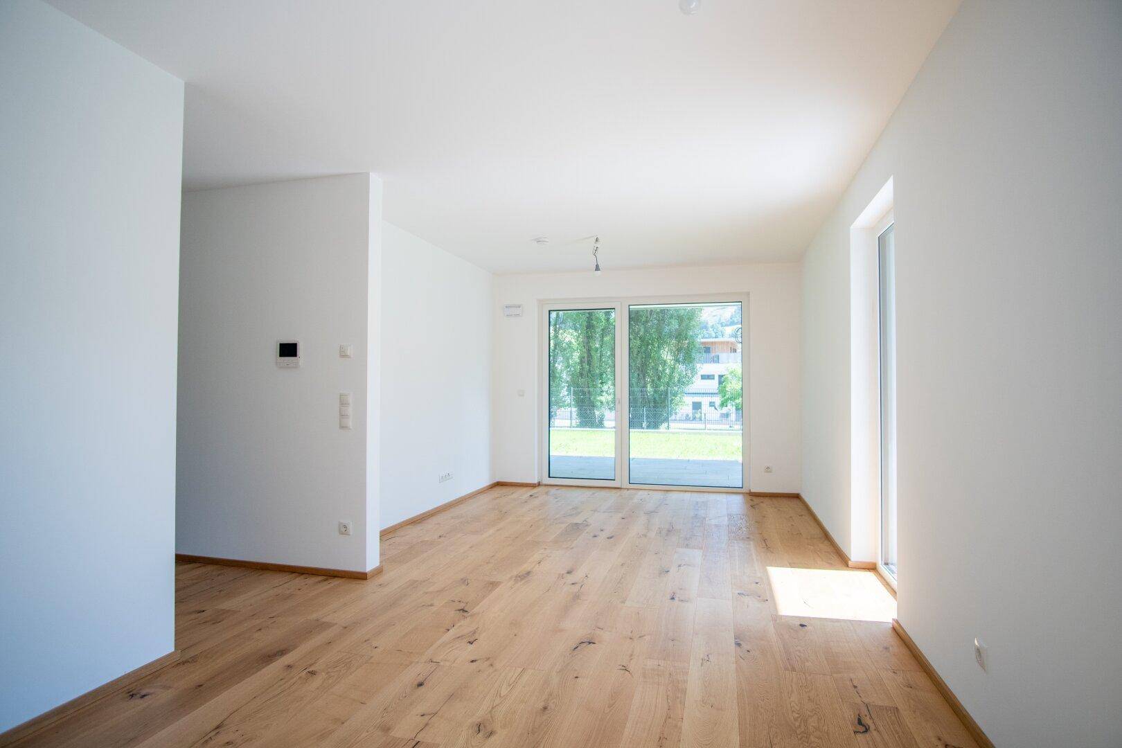 Leeres Wohnzimmer mit Holzboden, großen Fenstern und viel Tageslicht.