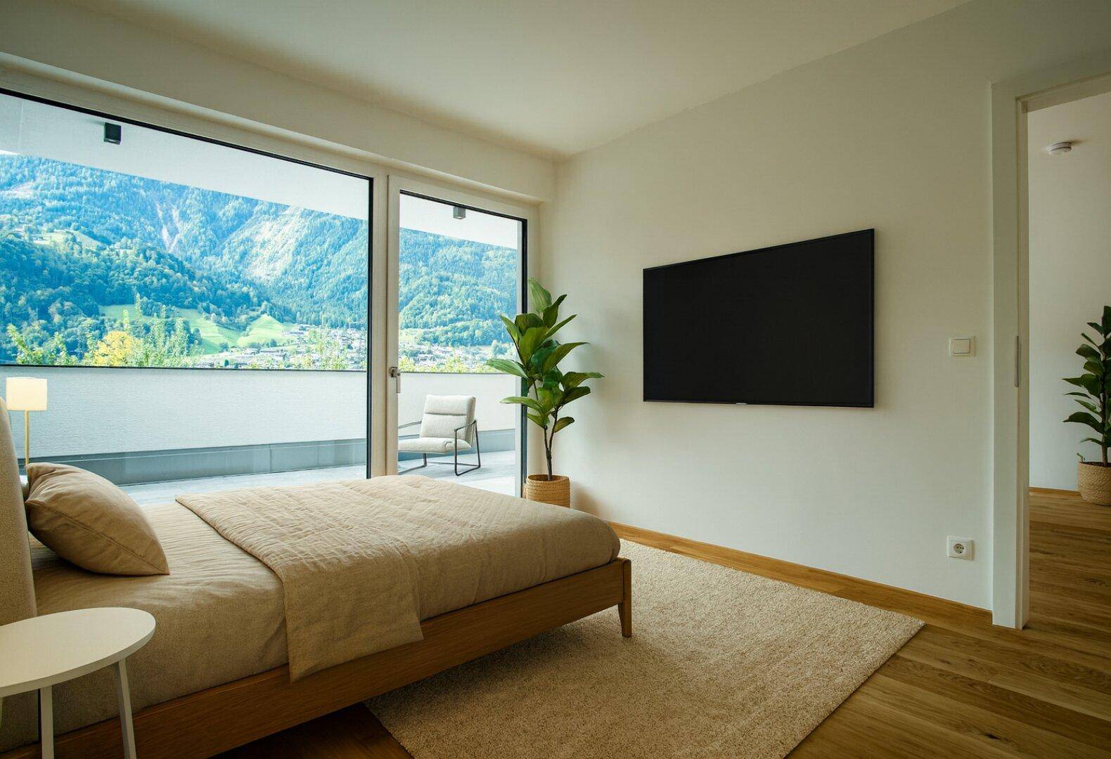 Schlafzimmer mit großem Bett, Holzboden und bodentiefen Fenstern mit direktem Zugang zur Terrasse und Bergblick.