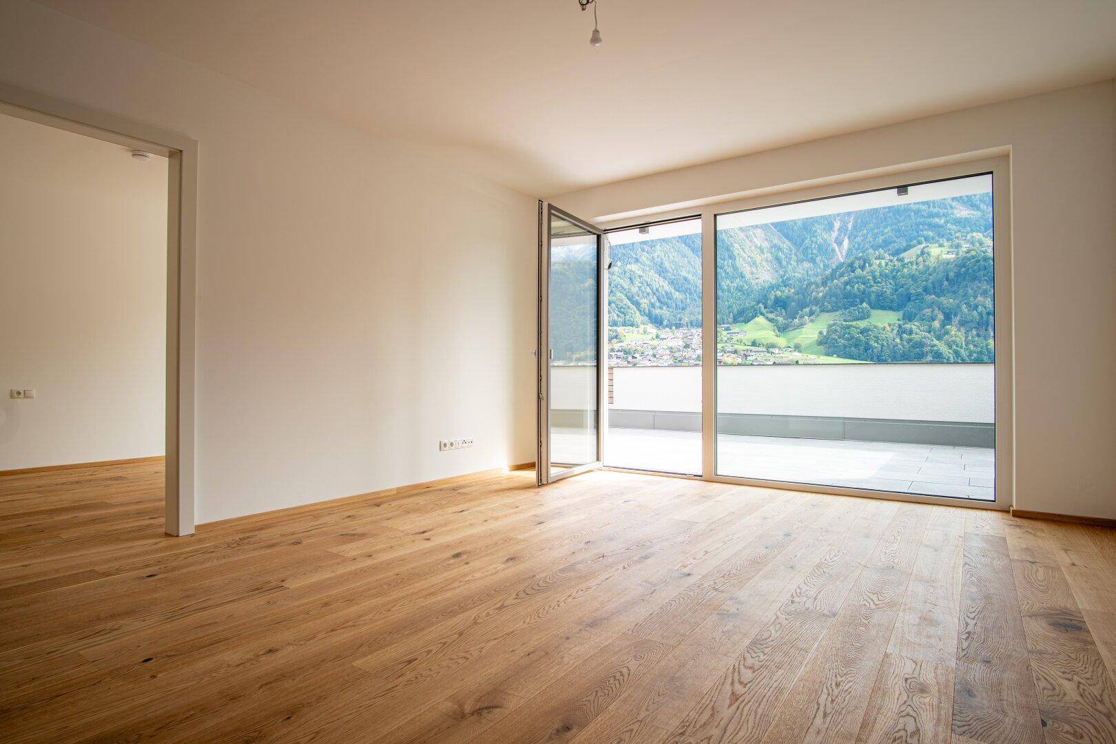 Heller, leerer Wohnraum mit Holzboden und großen Fenstern mit Zugang zu einer Terrasse und Bergblick.
